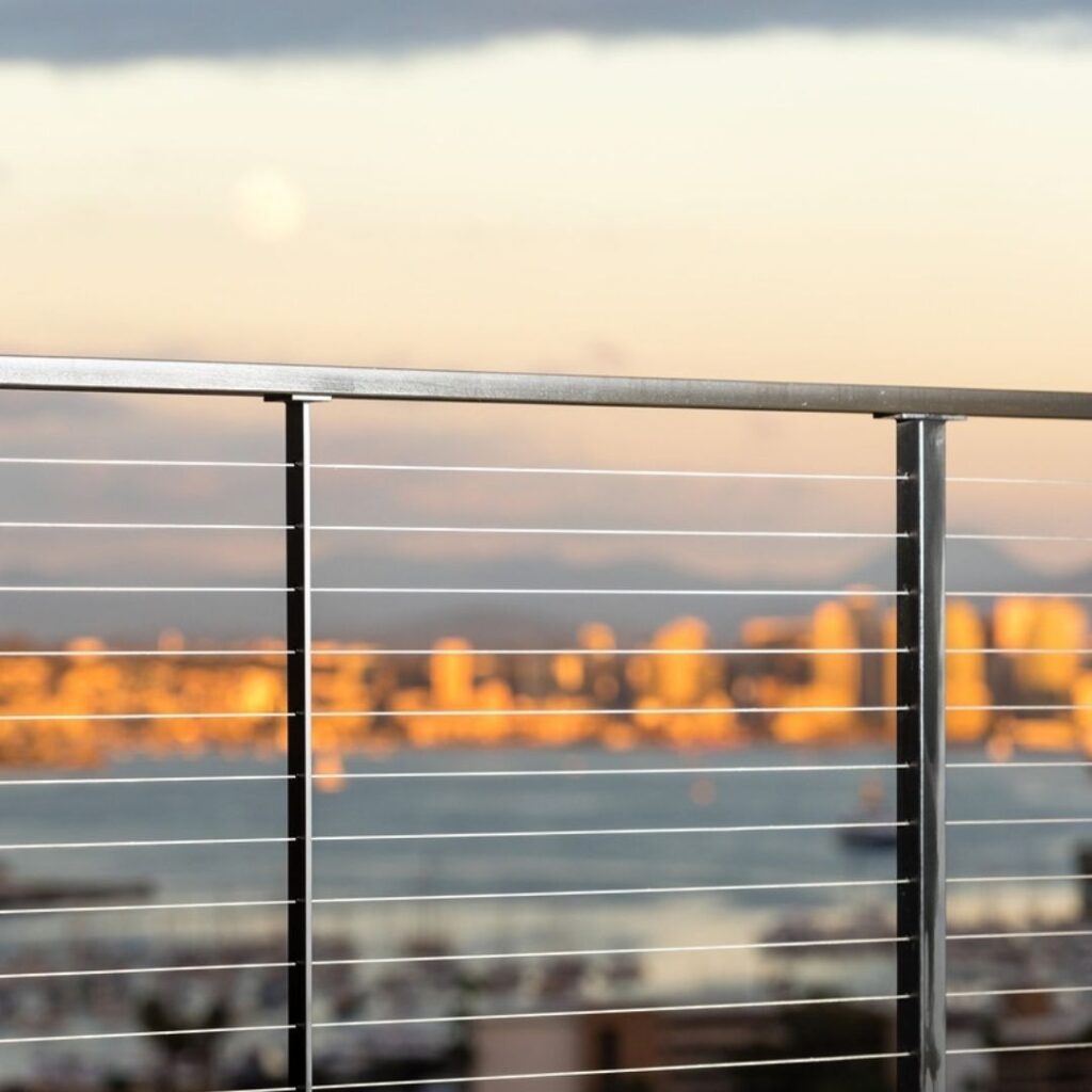 View from a balcony at sunset with a metal railing in the foreground, warm city lights and distant buildings blurred in the background, and a cloudy sky fading to pale orange near the horizon.