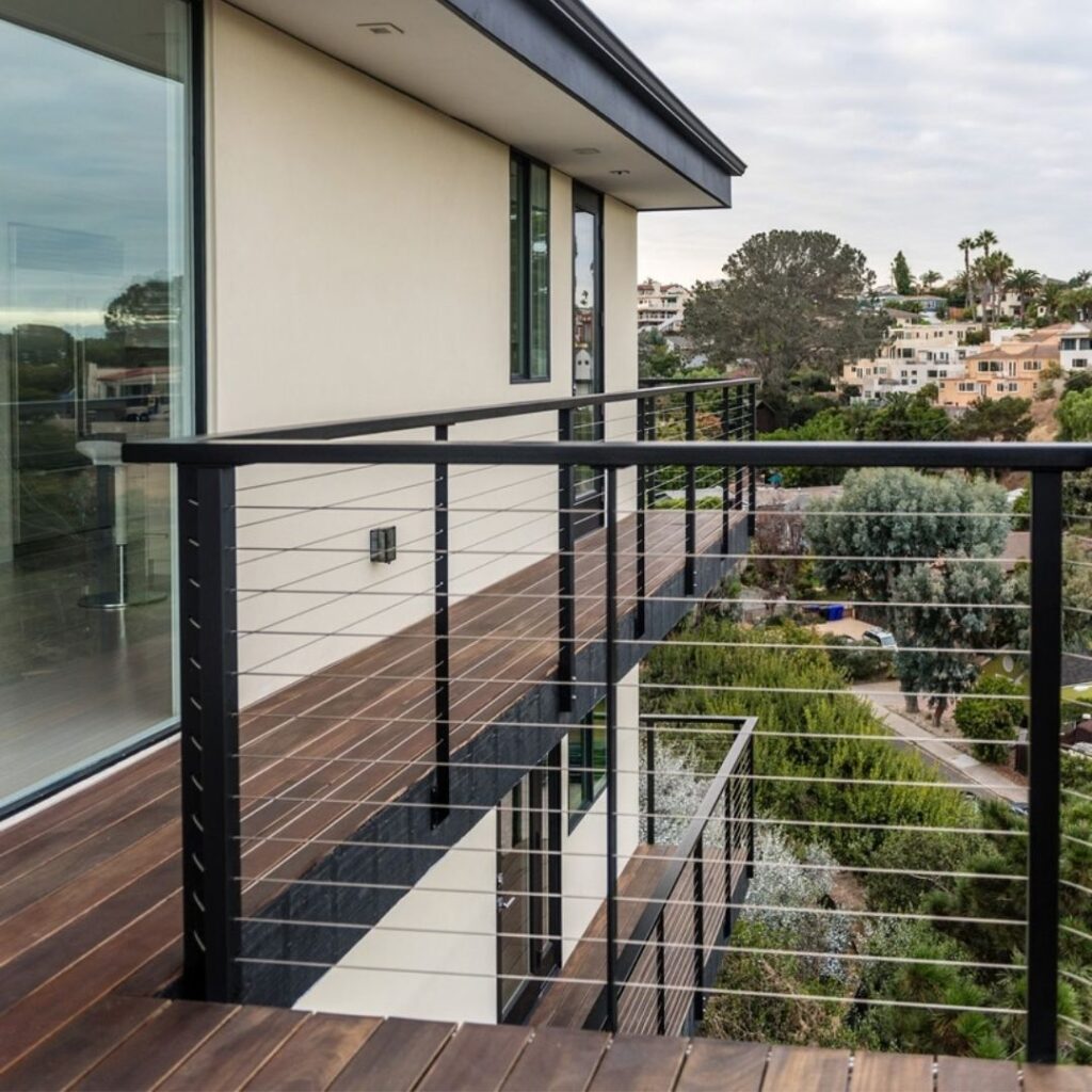 Modern building facade with large glass windows and a covered balcony, featuring dark metal railings and a view over a landscaped area and nearby hillside homes under a cloudy sky.