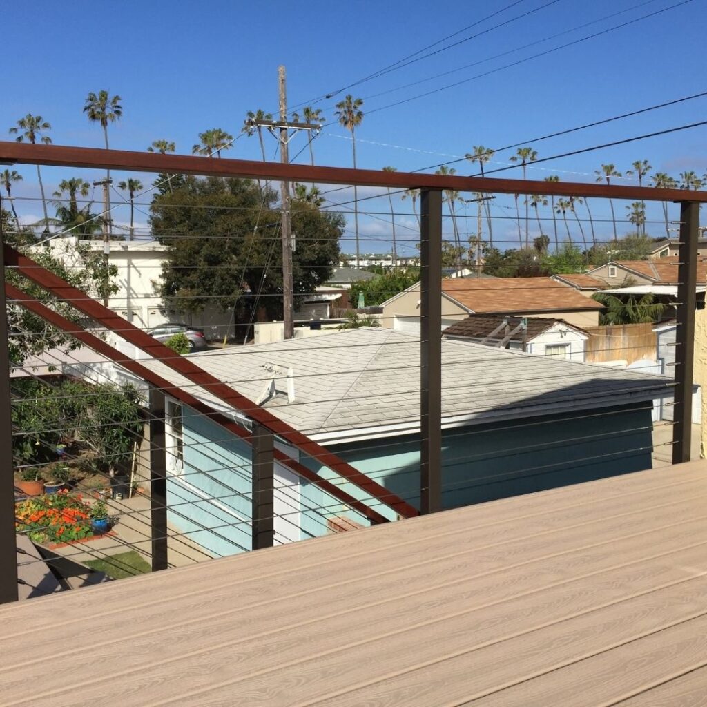 Outdoor view from a balcony with wooden railing and diagonal red-and-white beams, looking over rooftops, palm trees, and a backyard with a blue tarp under a clear blue sky.