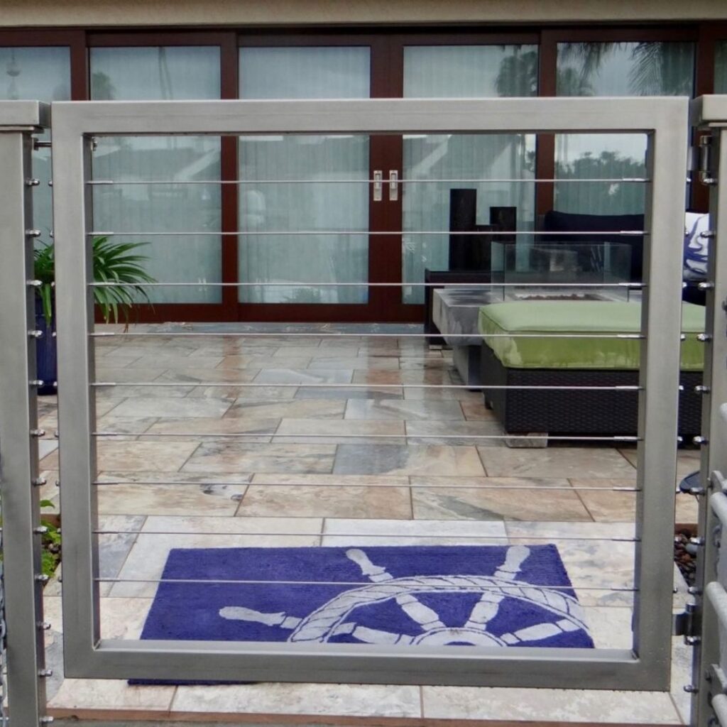 Glass patio door framed in dark metal, opening to a tiled courtyard with a blue-and-white doormat, a raised planter with green plants, and reflections of nearby trees in the windows.