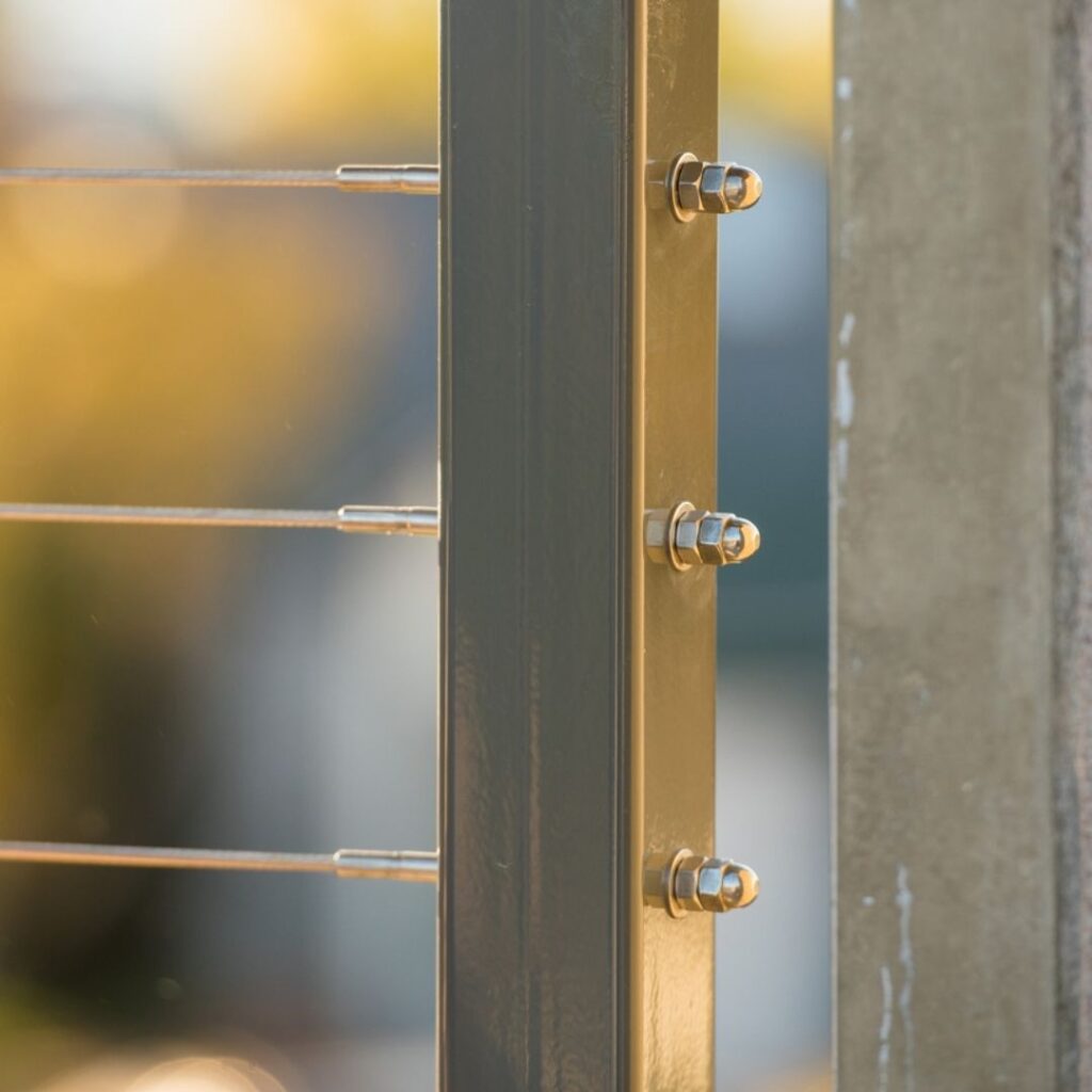 Close-up of a dark metal structural post with three bolts and a horizontal steel frame behind it, set beside a concrete wall.