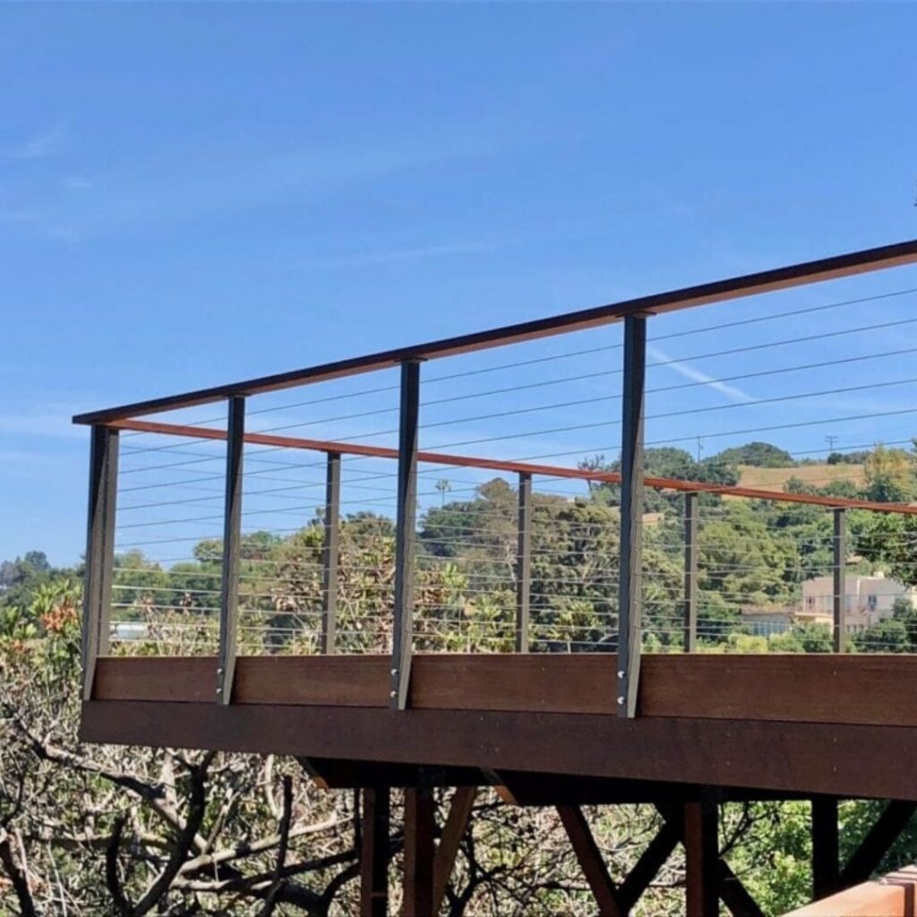 Outdoor view from a balcony with a rust-brown railing and metal posts, blue sky overhead, and green hills and buildings in the distance.